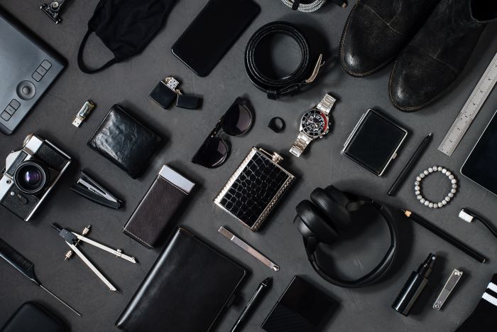 Assorted men's accessories including a watch, wallet, belt, camera, headphones, shoes, and electronic devices arranged on a dark background.